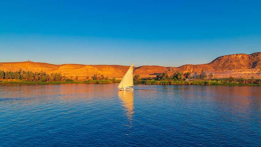 A sailboat with a white sail glides on a calm blue river against a backdrop of sunlit, golden desert hills under a clear, vibrant blue sky.