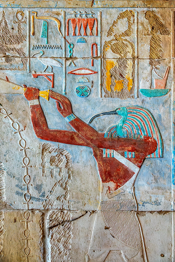 Ancient Egyptian relief depicts a red-skinned figure with a bird's head, possibly Thoth. Hieroglyphs above add historical context, colors vibrant.