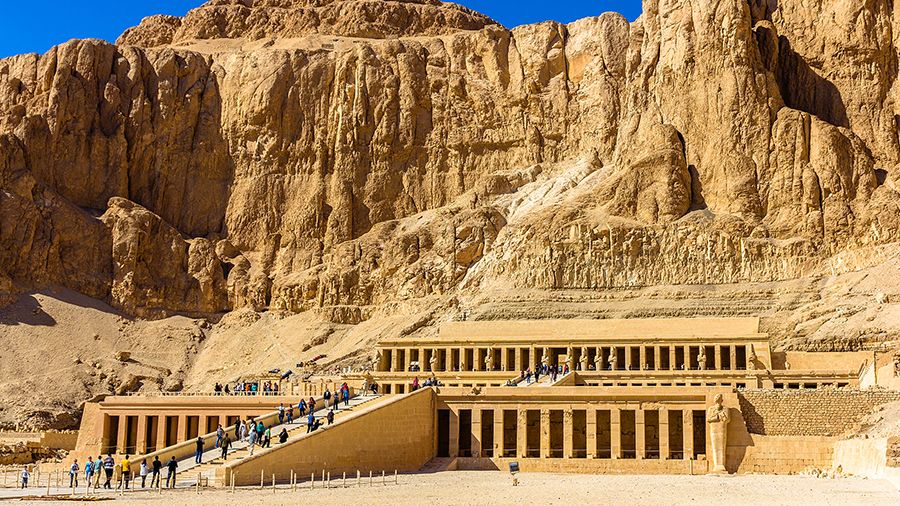 Ancient Egyptian temple with terraces and columns set against a rugged cliff under a bright blue sky. Tourists explore the historic site, conveying awe.