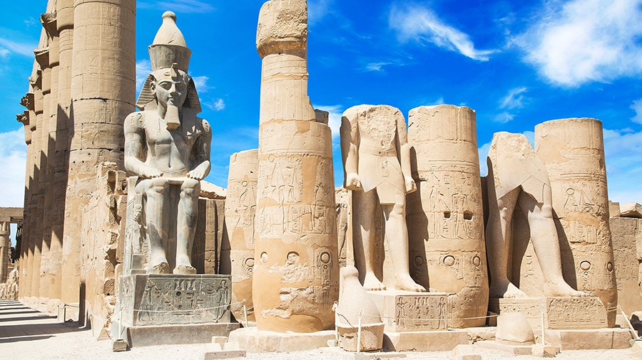 Ancient Egyptian statues and columns at Luxor Temple under a bright blue sky. The large seated statue is prominent, conveying a sense of grandeur.