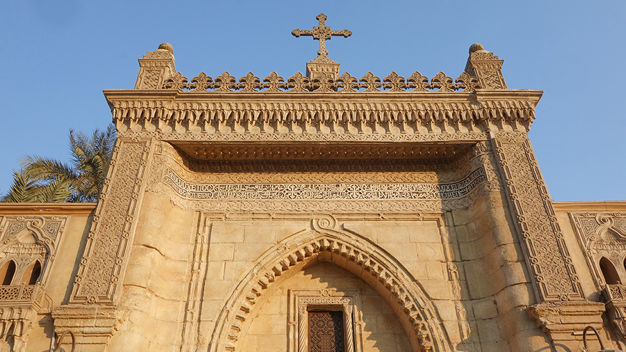 Ornate stone church facade with intricate carvings and an arched window, topped by a cross. Palm leaves peek from the left, under a clear blue sky.
