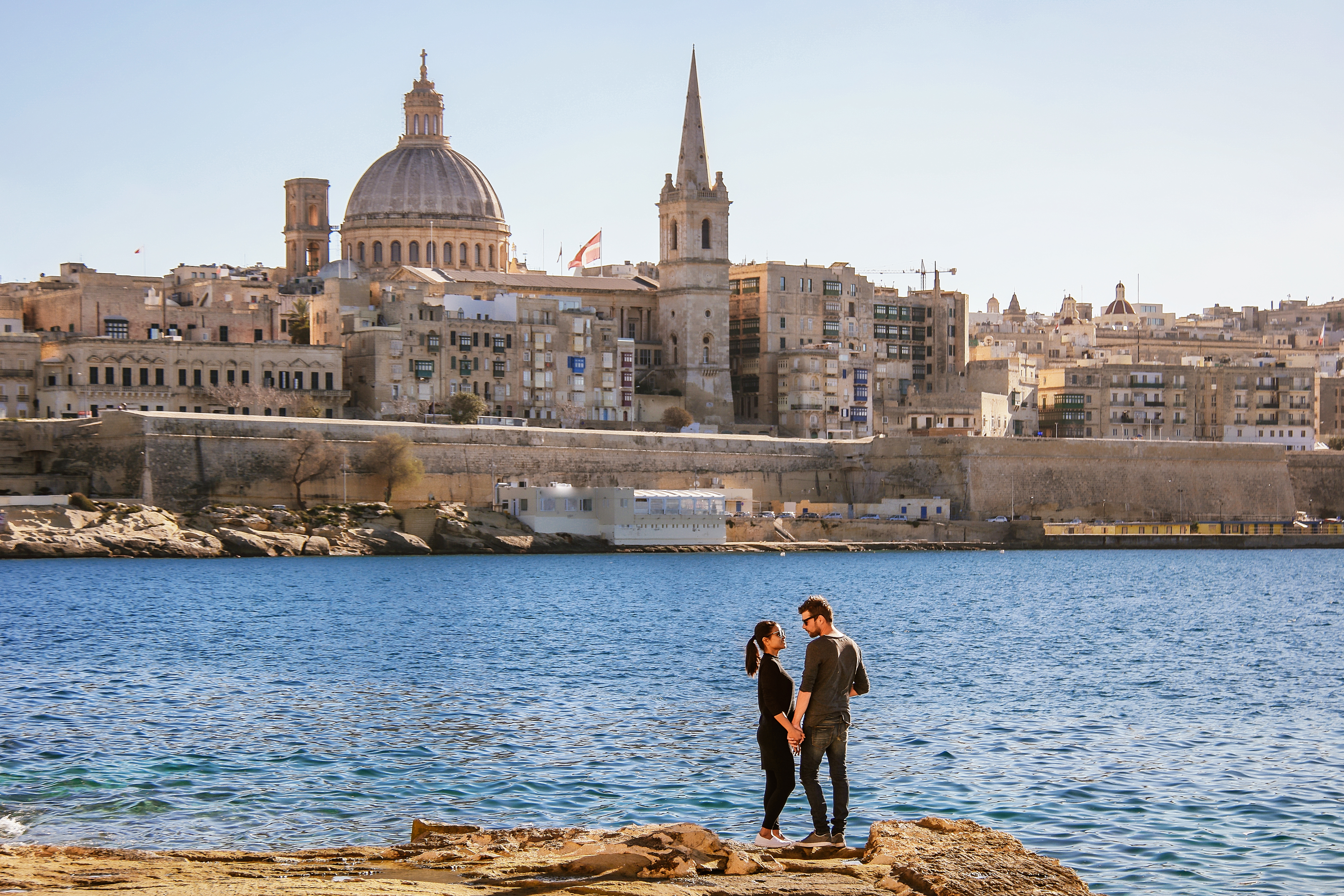 People standing by the sea with Valletta skyline and St. Paul’s Cathedral in the background.