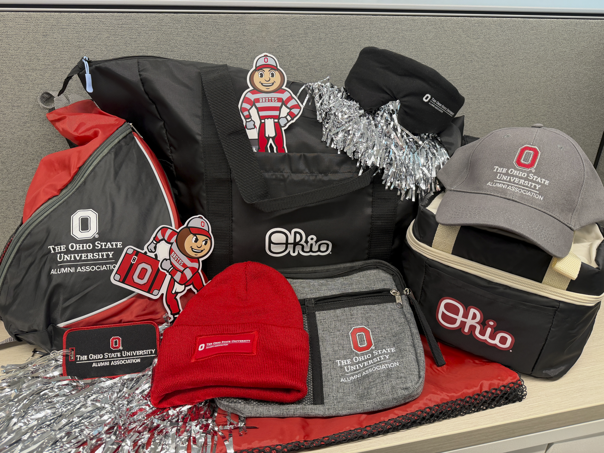 A collection of Ohio State University Alumni Association branded items arranged on a desk, including black and red duffel bags, a gray cooler bag, a gray baseball cap, a red knit beanie, a gray zip pouch, silver pom-poms, and Brutus Buckeye stickers and cutouts, with “Ohio” and Ohio State logos visible on several items.