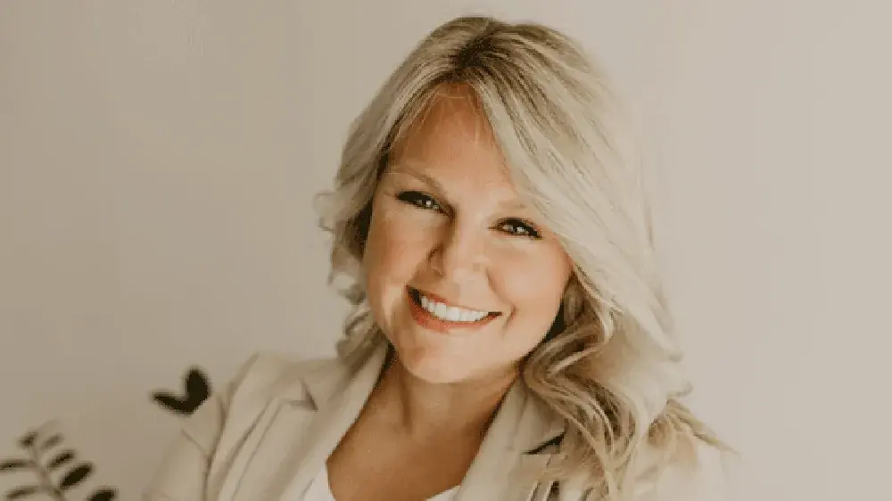 Aliza Litke wearing a white blouse and kight jacket in front of a tan while smiling