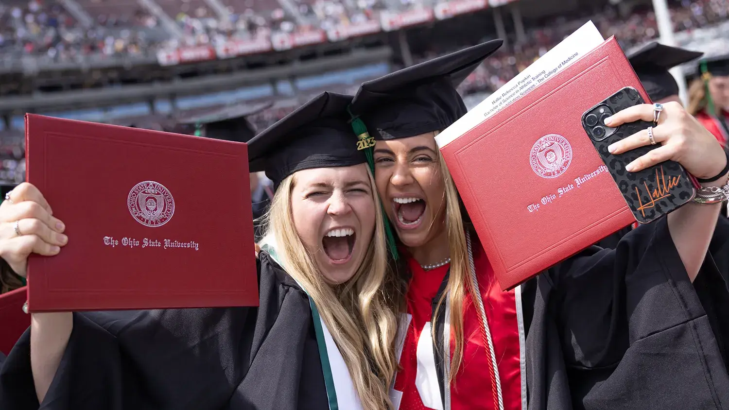 two women smile while holding their college diplomas