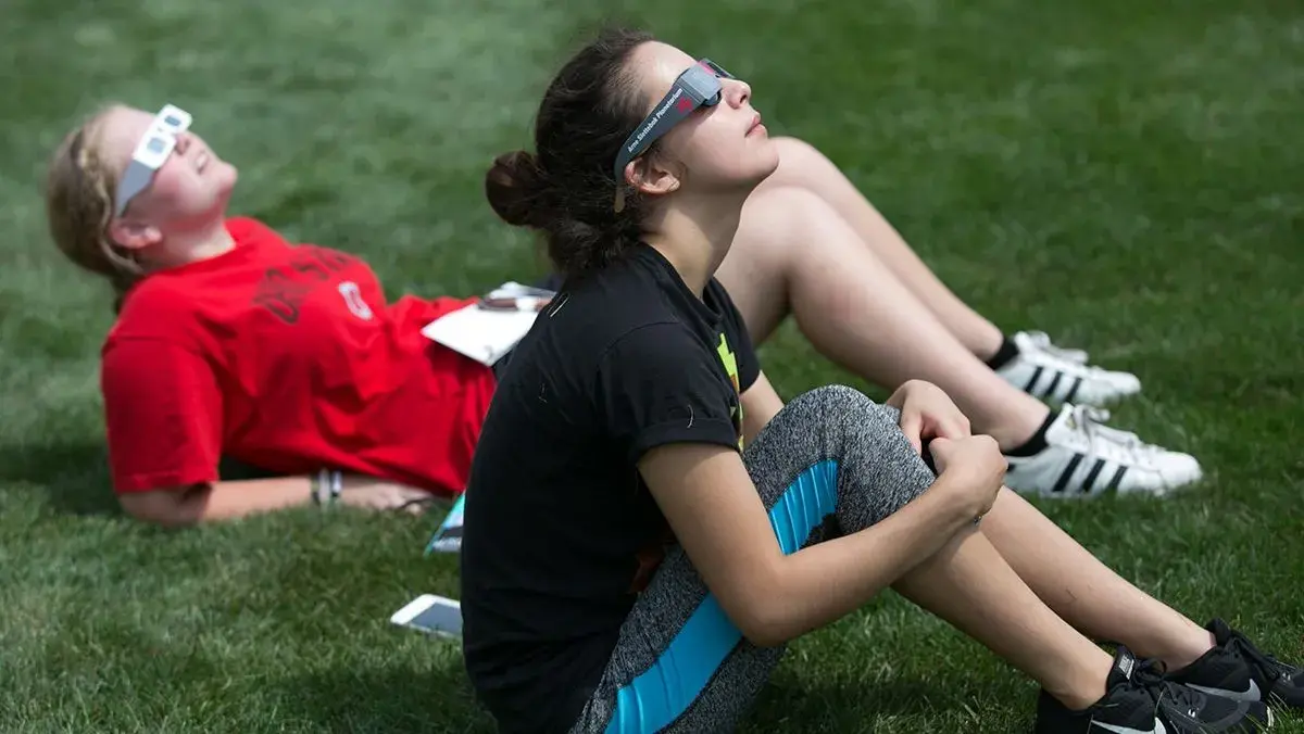 two people sitting on grass wearing solar eclipse glasses