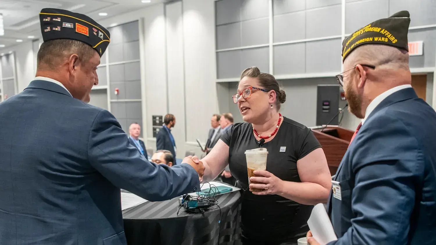 At a table on a small stage, Gretchen Klingler, a white woman with a style reminiscent of the 1940s, leans across a table to shake hands with a white man wearing an American Legion uniform cap and a suit. They’re making eye contact, and next to them smiling is a white man in a suit, this one with a VFW uniform cap