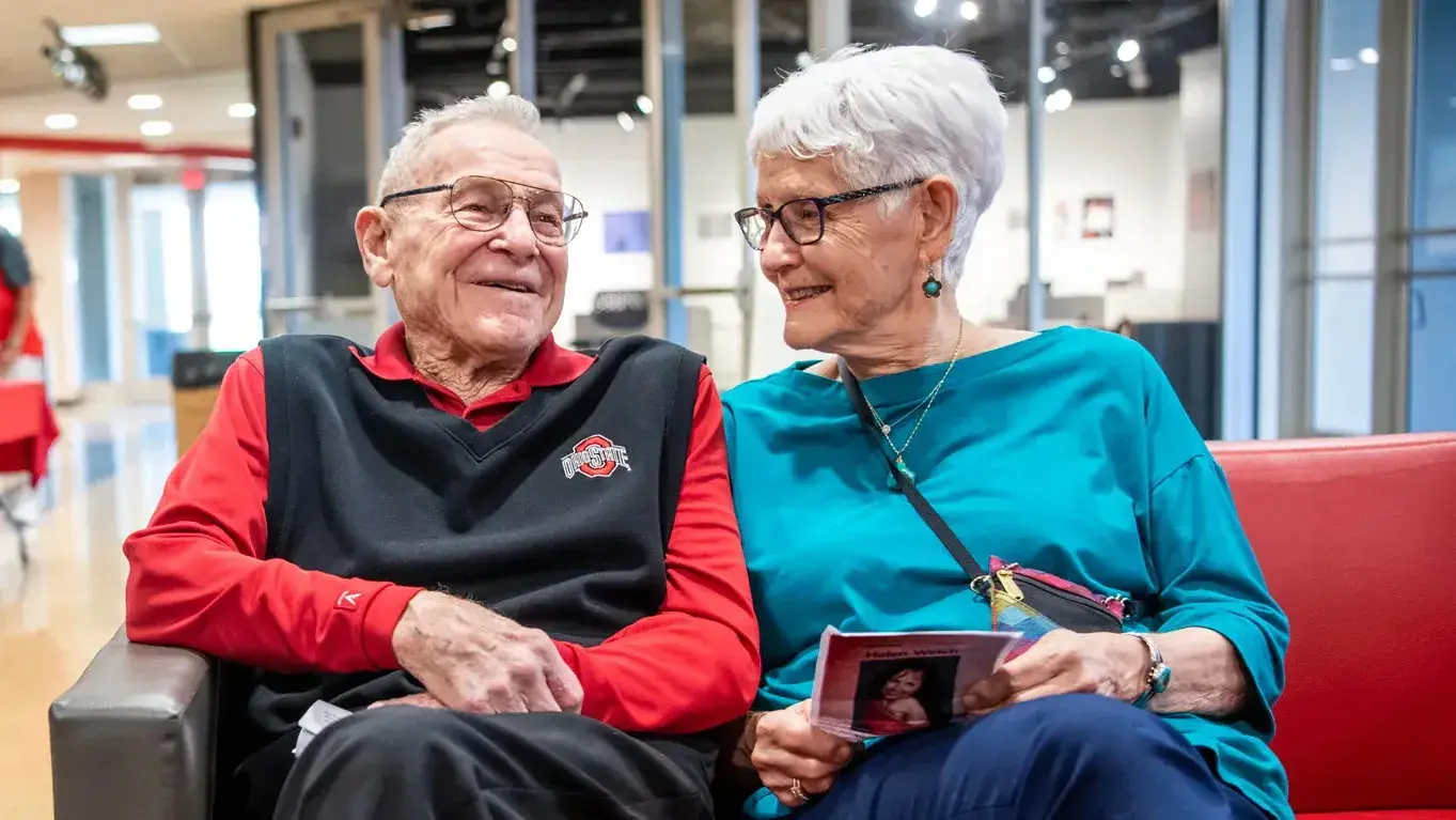 A white-haired couple sit together on a couch, smiling as they chat and lean against each other.