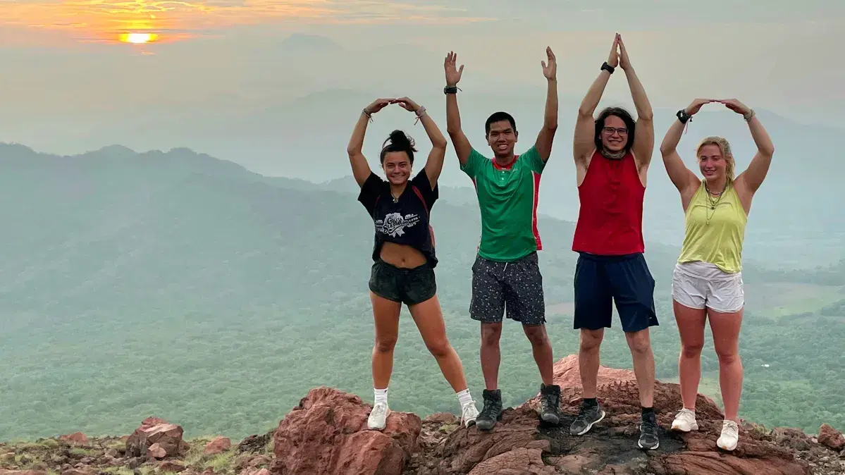 Four people pose on a rocky ledge with mountains behind them, forming 'O-H-I-O' with their arms at sunrise or sunset.