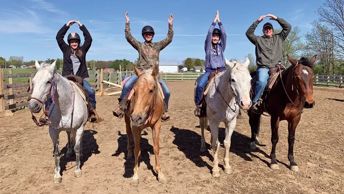 Four people on horses form 'O-H-I-O' with their arms in an outdoor arena. All are wearing helmets.