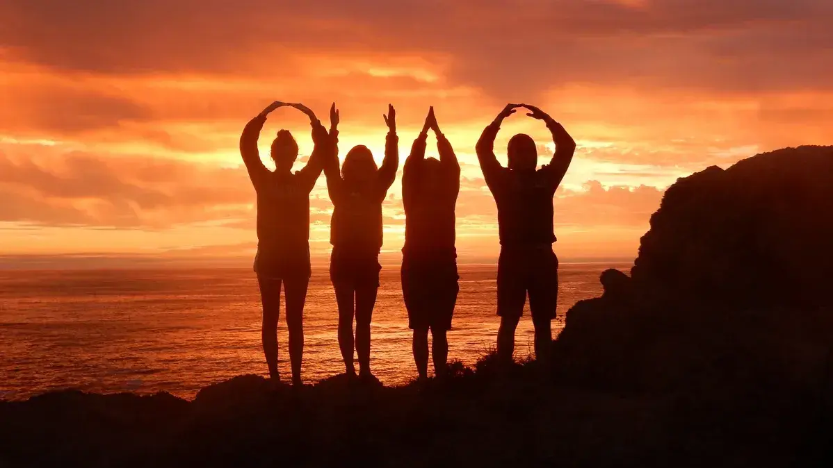 Four people stand on a rocky shore at sunset, forming ‘OHIO’ with their arms. The ocean and colorful sky are in the background.