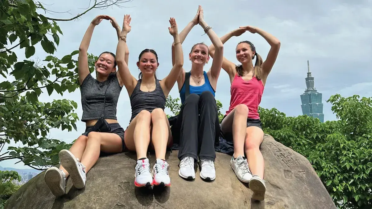 Four people sit on a rock, forming the word 'OHIO' with their hands. Trees and a tall building are in the background.