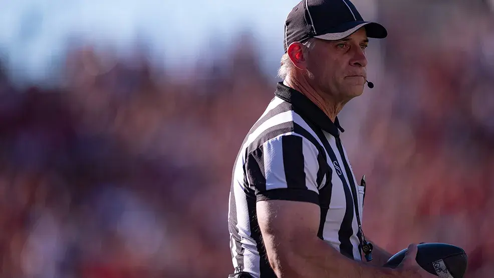 A football referee holds the ball and makes a thoughtful face as he watches something out of the photo frame. He wears a black and white striped referee’s shirt, a ballcap and a headset that leaves a small mic in front of his mouth to communicate calls.