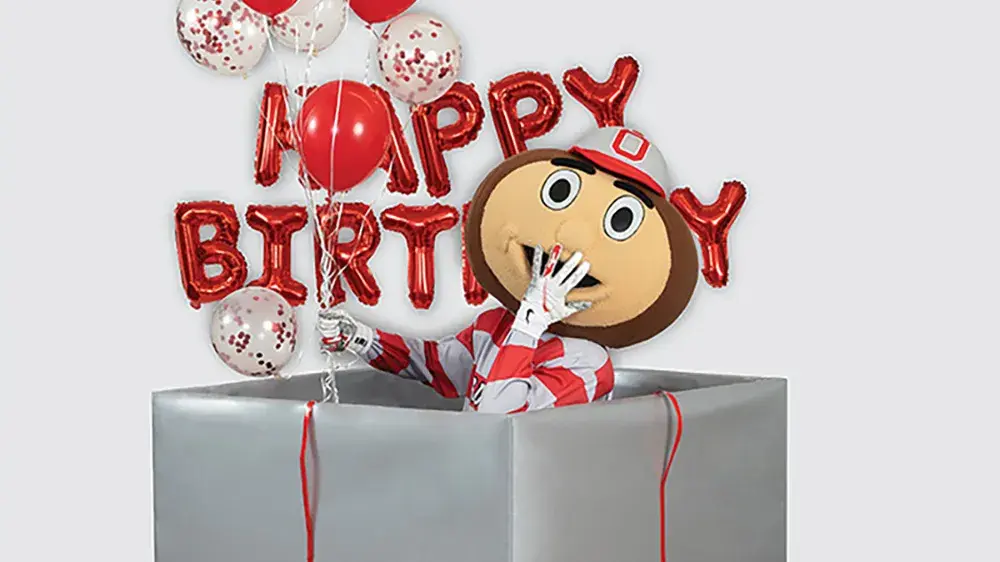 Ohio State's mascot, Brutus Buckeye, holds a bouquet of balloons and giggles with a hand over his mouth as he pops out of a gigantic gift box