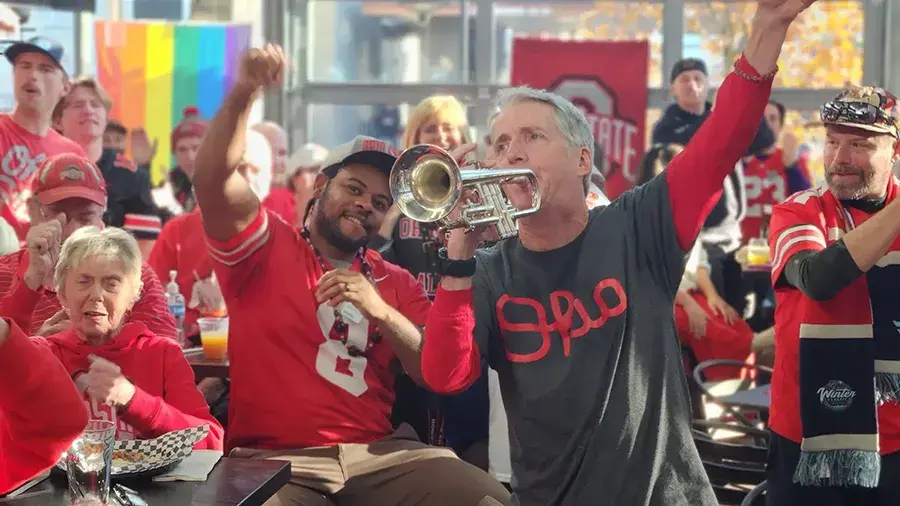 Man in gray Script Ohio shirt playing the trumpet around a group of poeple.
