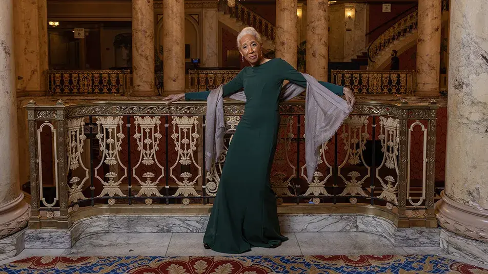 Dianne McIntyre, a slender and stylish black woman, poses along an ornate railing in a historical theatre with big marble columns and intricate detailing. 