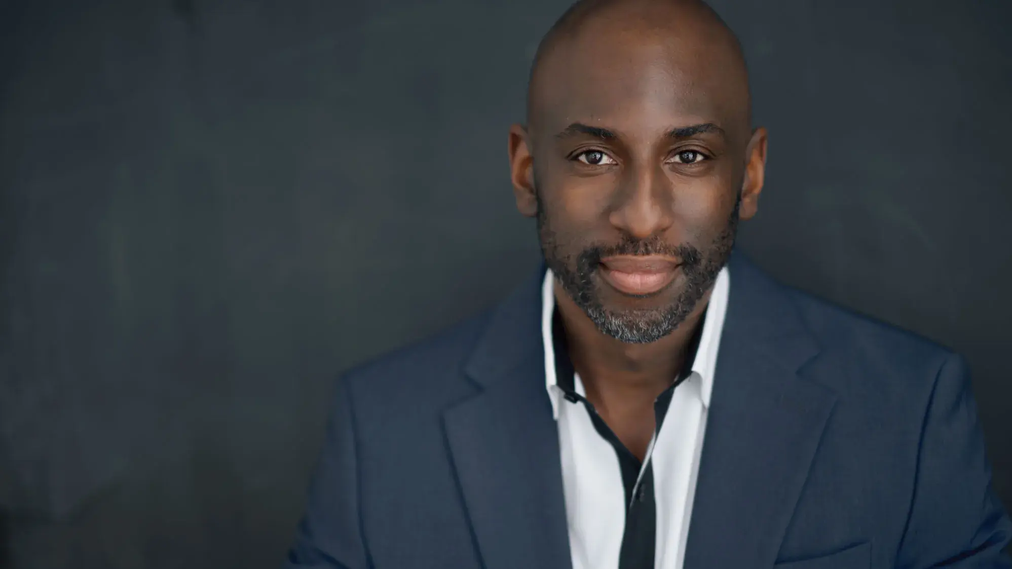 Rashad Chambers poses for a portrait wearing a suit and button down shirt. A young-ish black man, he appears stylish, calm, confident and proud.
