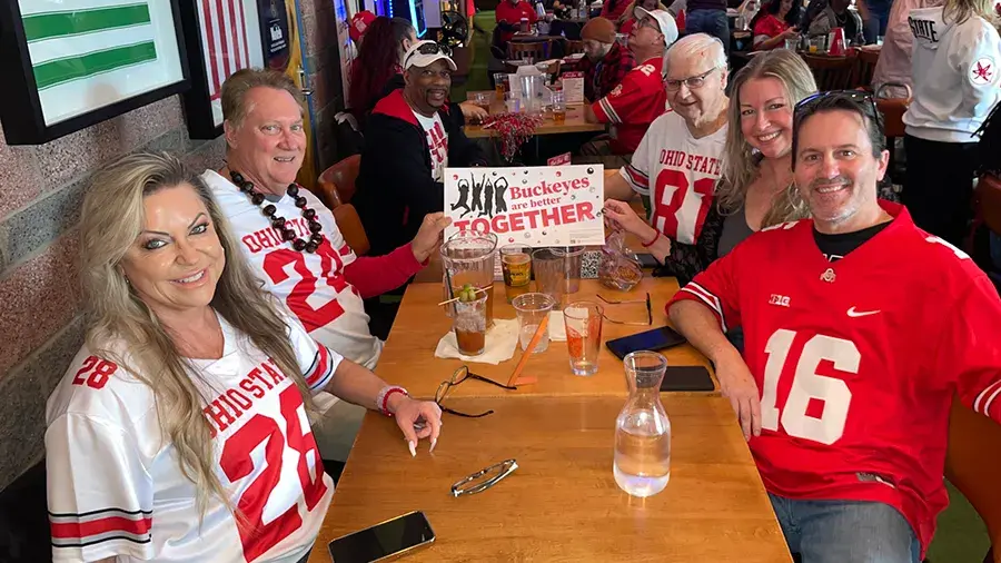 Group of people wearing Ohio State Buckeyes jerseys seated at a wooden table in a sports bar. One person holds a sign that reads: “Buckeyes are better TOGETHER.” The background shows more fans in similar attire and sports-themed decor.