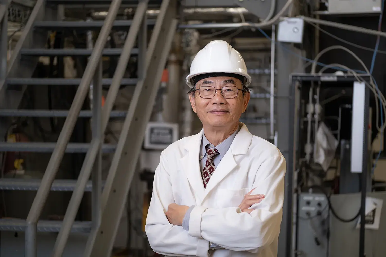Liang-Shih Fan seems quietly proud and confident, as he poses for this portrait. He wears round glasses and a white lab coat and hard hat over his suit. 