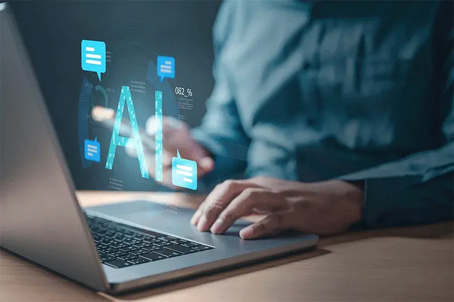 A person sitting at a desk typing on a laptop computer with a digital illustration of the acronym AI floating above the computer in blue font
