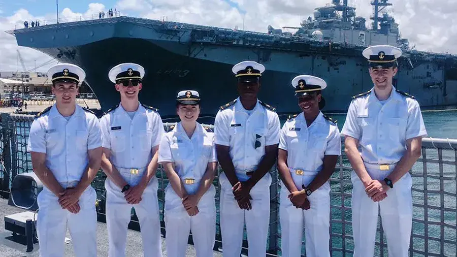 Navy members standing in their white uniforms with a naval vessel in the background.