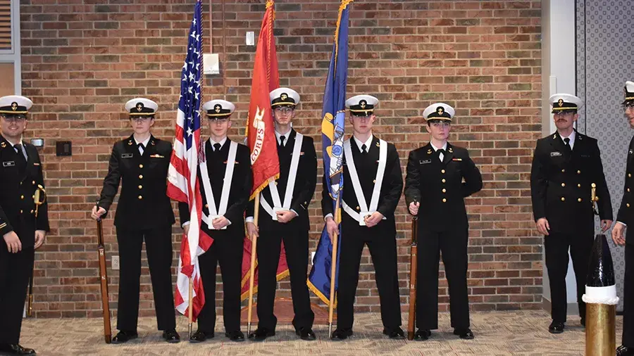 Cadets in uniform standing in front of a brick wall indoors holding flags. 