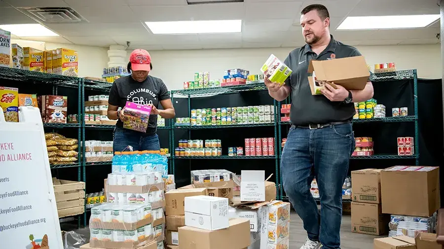 Two people organize canned goods and boxed food items in a storage room with shelves stocked with pantry supplies and stacks of boxes on the floor.