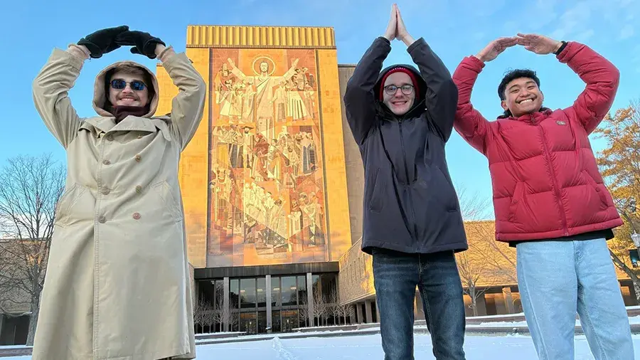 Three people standing in the snow posed with their arms raised to form letters O-H-I-O in front of a tall building with a large mural.