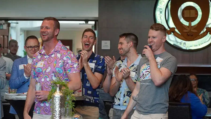  A group of people stand together indoors, some clapping and smiling, in a casual social setting with colorful shirts and a university-style emblem on the wall behind them.
