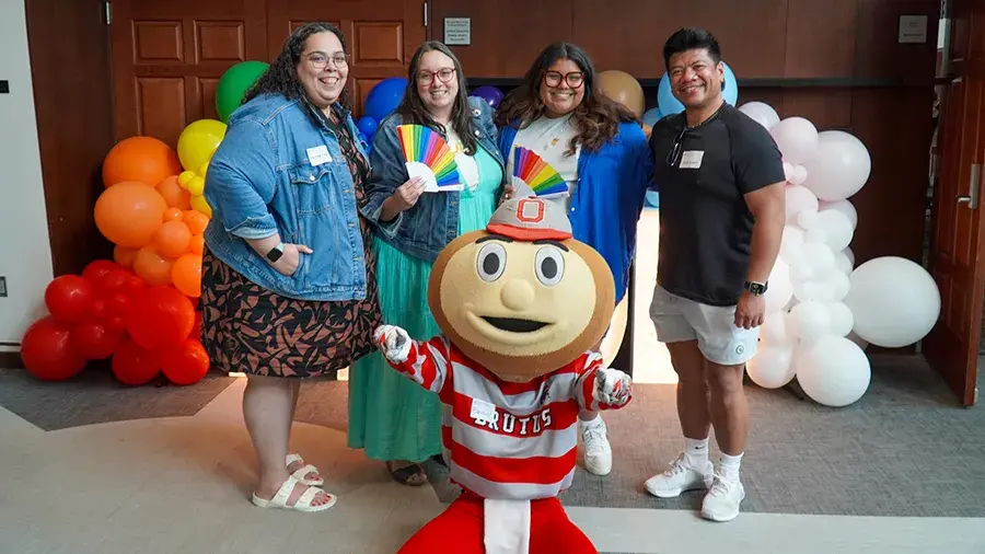 Brutus Buckeye kneels in front of four people standing together. Behind them is a colorful balloon display arranged in a rainbow pattern.