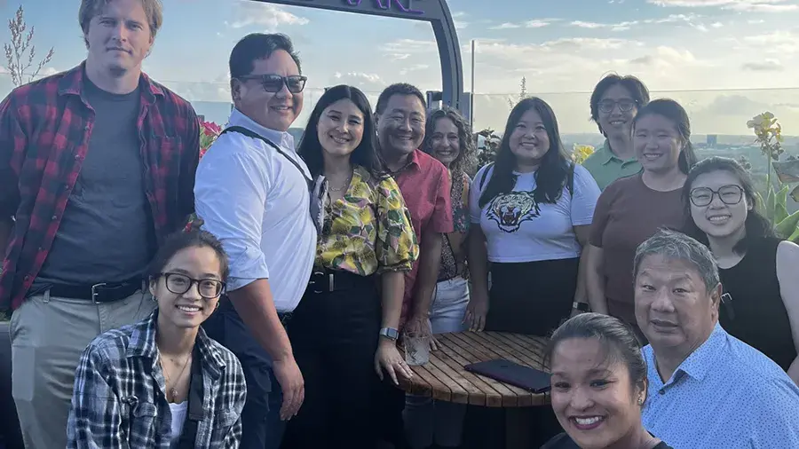 A group of people standing and sitting together around a small outdoor table on a rooftop patio with plants and a city skyline in the background.
