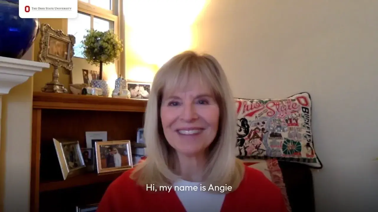 A person sits indoors in a home office setting, wearing a red top, with bookshelves, framed photos and decorative items behind. A pillow with Ohio State-themed designs is on the chair, and the video subtitle reads, “Hi, my name is Angie.”