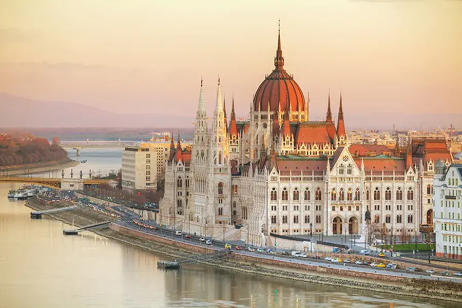 A large, ornate riverside parliament building with a red dome and spires at sunset.