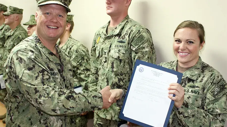 U.S. Navy personnel in uniform participate in a recognition ceremony, with one service member holding a certificate and another shaking hands.