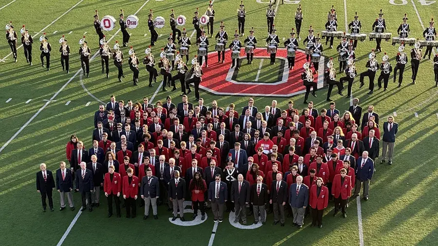 A large group of people in suits and red jackets standing on a football field in front of a marching band.
