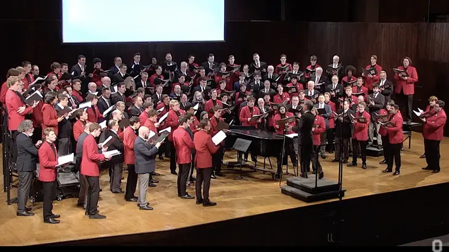 A large choir of people in red blazers and dark suits performs on a concert stage while a conductor directs them.
