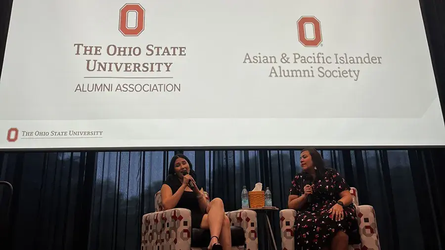 Two speakers seated on a stage having a discussion beneath a large screen displaying logos and text for The Ohio State University Alumni Association and the Asian & Pacific Islander Alumni Society.