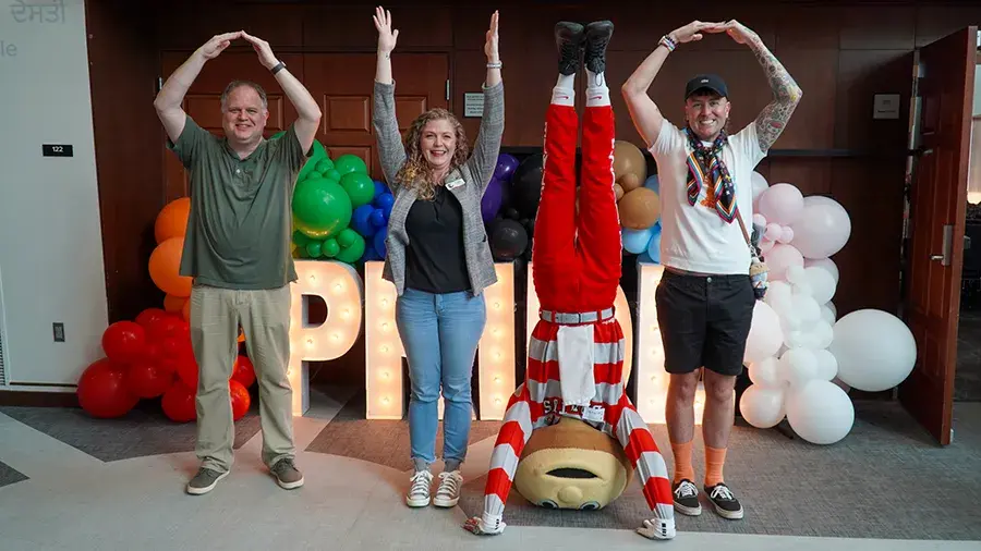 Four people stand in front of colorful balloon decorations and large light-up letters forming the word “PRIDE,” each posing to form the O‑H‑I‑O shapes, with Brutus Buckeye doing a handstand to make the “I.”