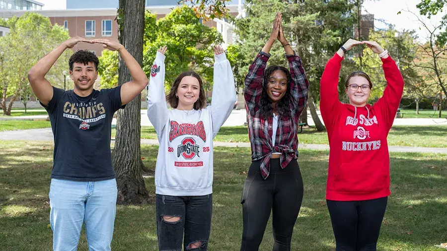 Four people standing outdoors on a campus lawn, each forming a different letter with their arms to spell “O-H-I-O,” all wearing Ohio State–themed clothing.