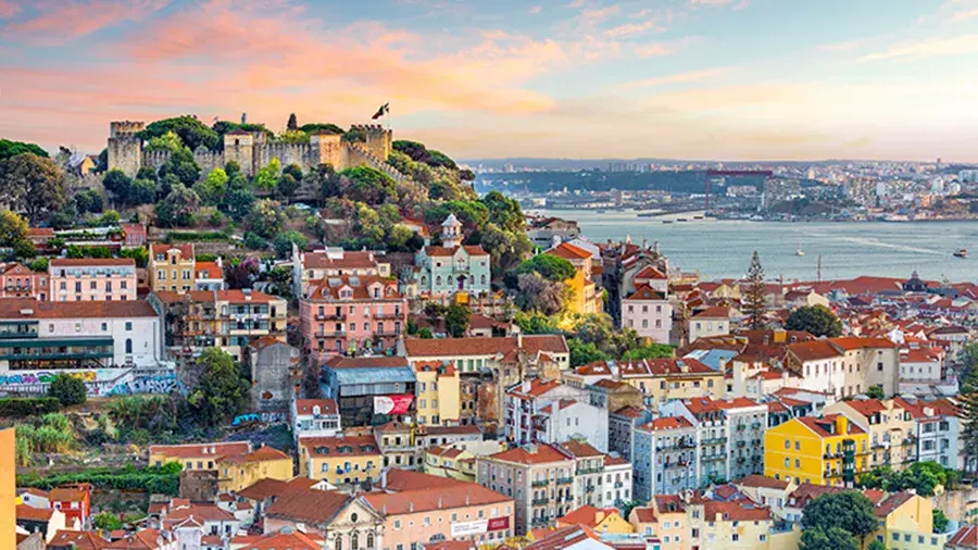 A colorful hillside view of the country side with red-roofed buildings, a historic castle on a hill, and a river in the background at sunset.