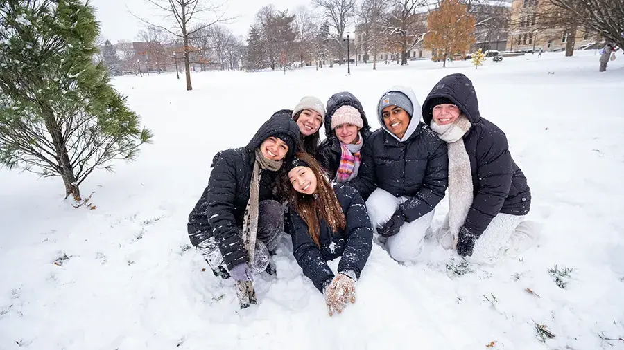 A group of bundled‑up people kneeling and sitting together in the snow on a snowy campus lawn, surrounded by trees and buildings in the background.