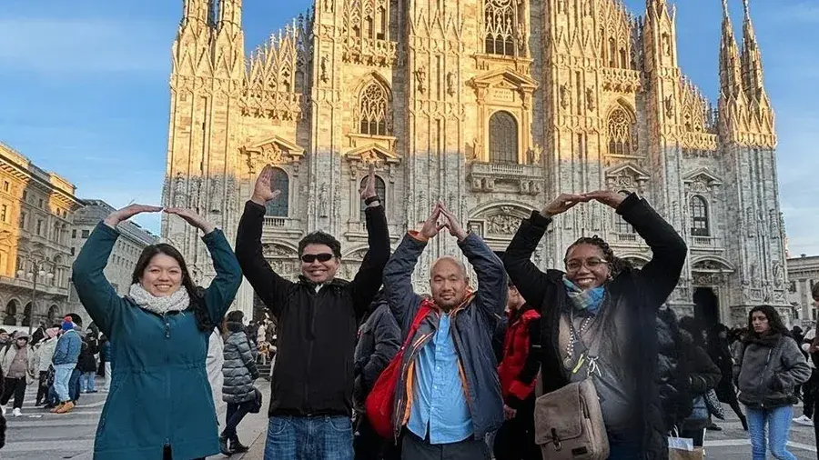 A group of four people stand in front of a large, ornate cathedral, each raising their arms to form letter shapes. The scene takes place in a busy public square with many other people in the background.