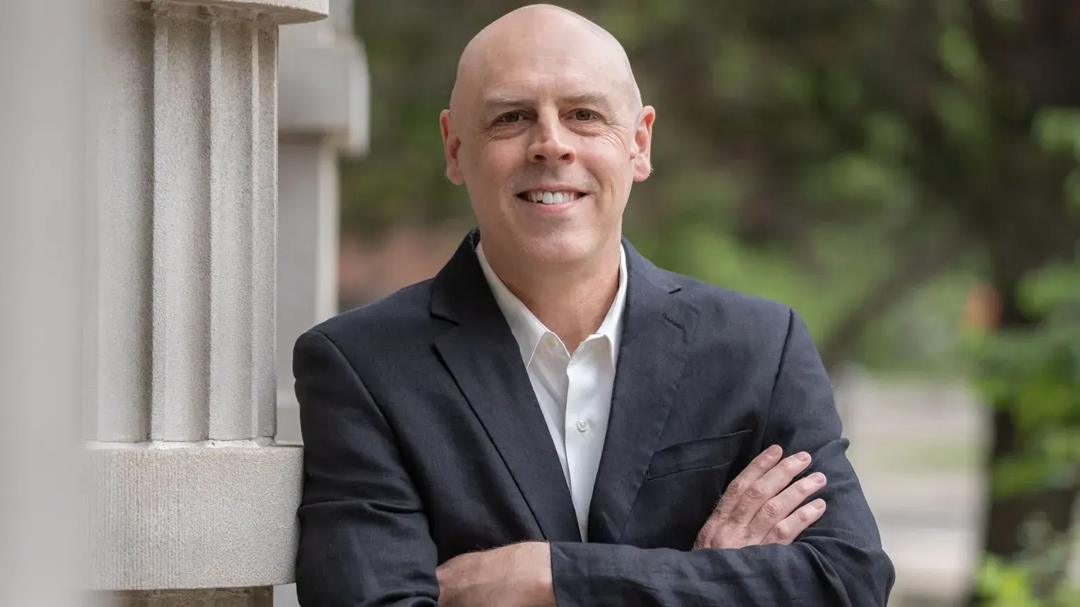 A professor smiles as he stands outdoors leaning against a column with arms crossed, wearing a collared shirt and suit jacket. He is a white man with a shaved head. Trees and a walkway appear in the background of the photo.