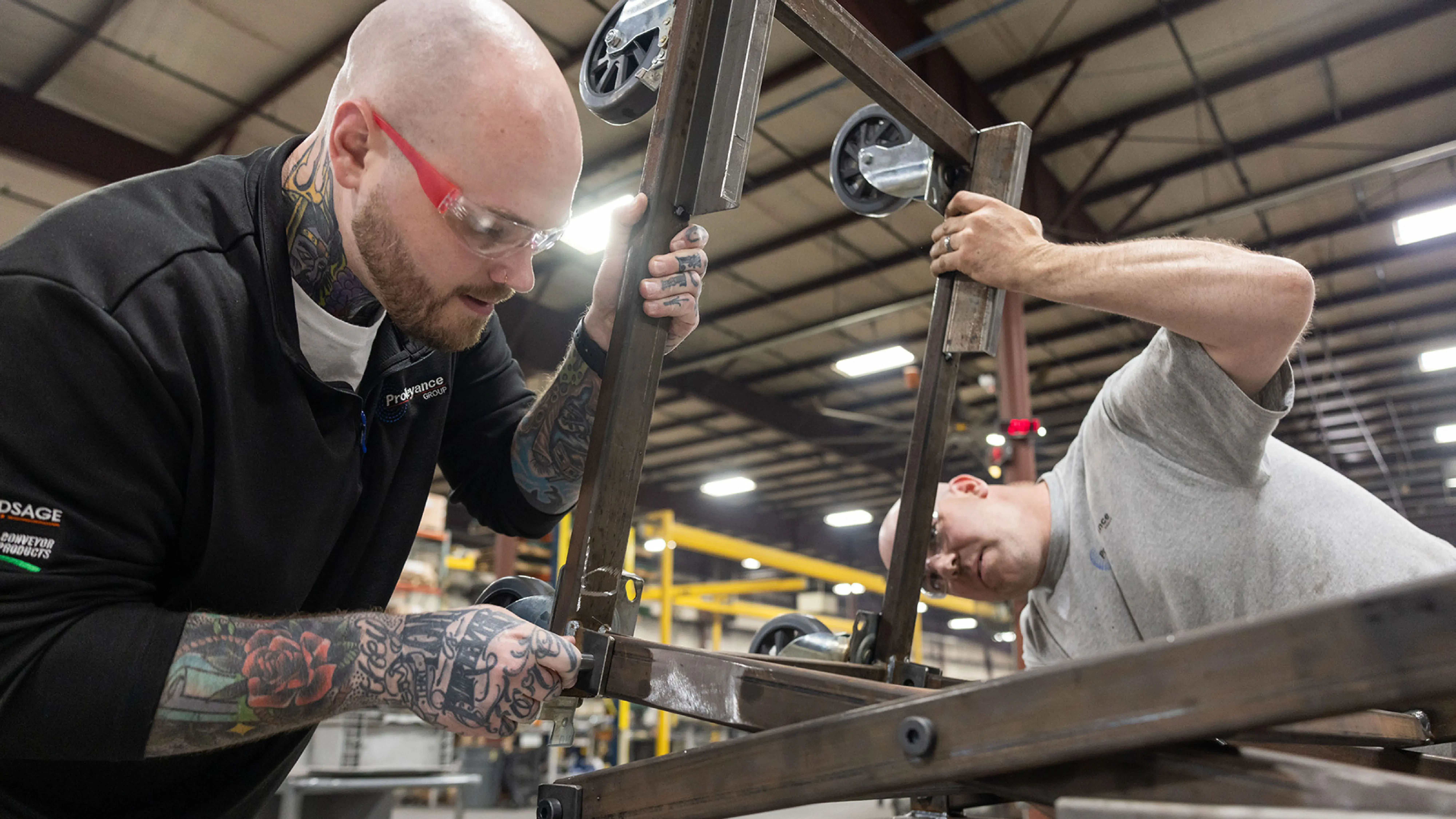 Two individuals are working together in an industrial workshop, assembling or adjusting a metal frame with caster wheels under bright overhead lighting. One person is holding the frame steady while the other tightens a component. The setting includes metal structures and equipment typical of a manufacturing environment.