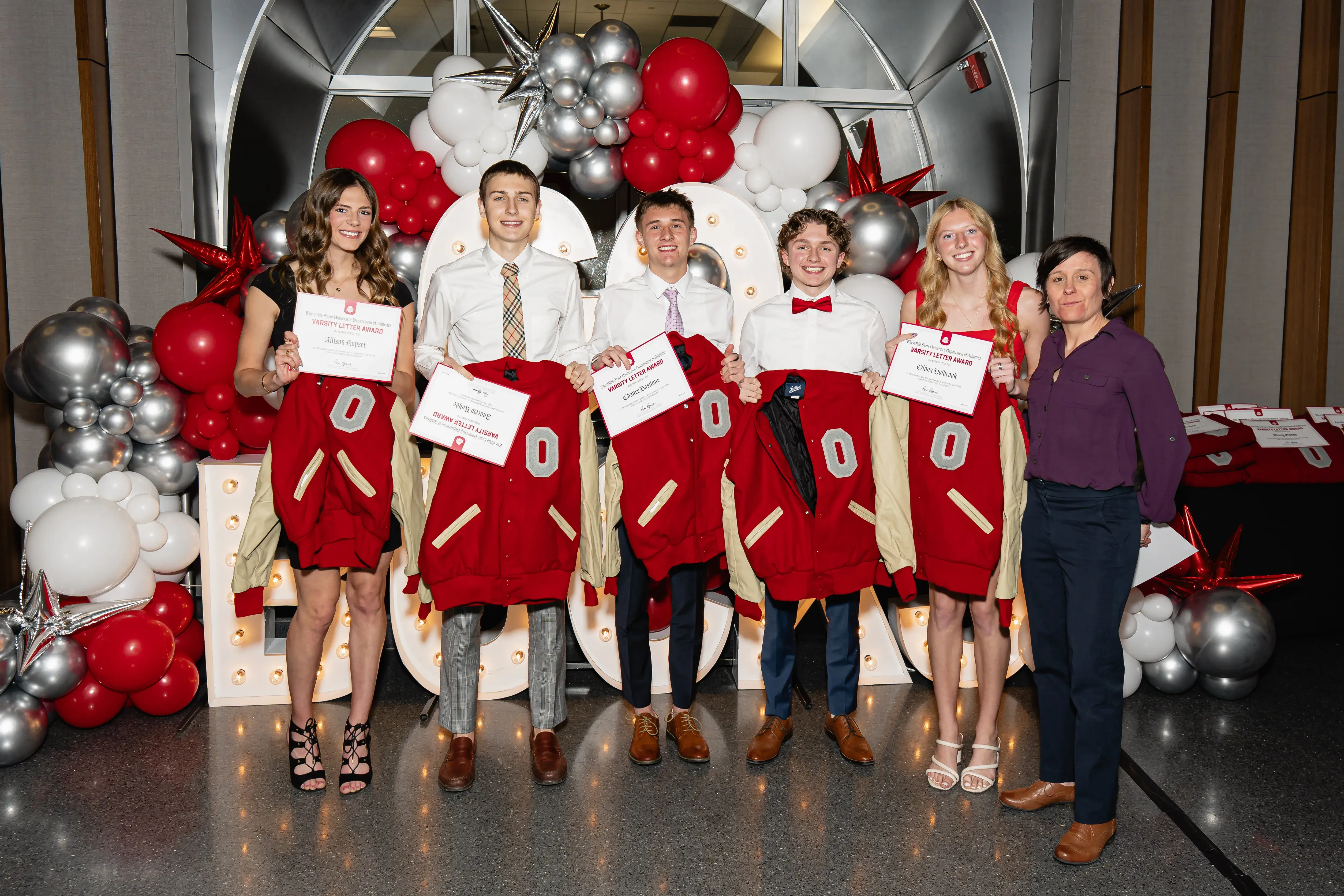 varsity O letter winner receivers holding their award and letter jacket together smiling infront of a decorative balloon structure 