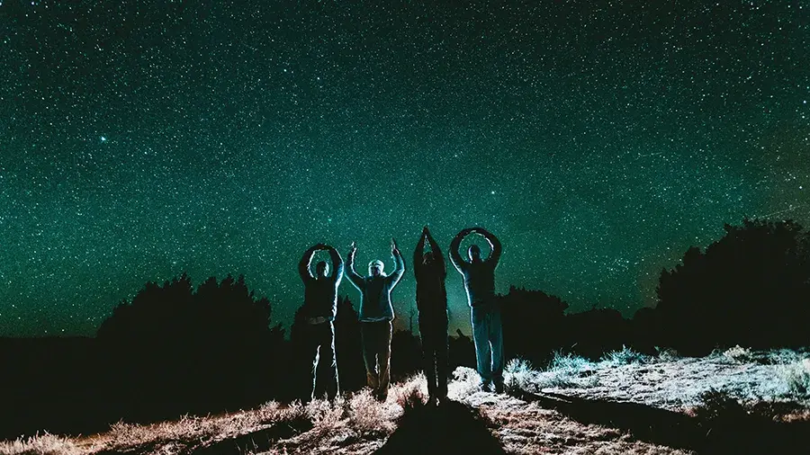 A group of four people standing on a hill at night, silhouetted against a star-filled sky, with their arms raised in different shapes.