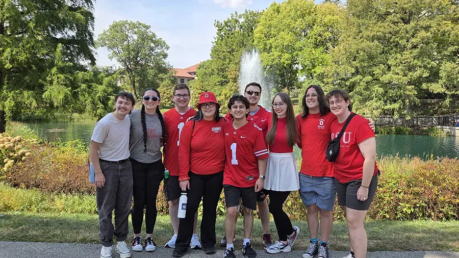 A group of people wearing matching red and neutral-colored clothing stand together outdoors in front of a pond with a large fountain, surrounded by green trees and landscaping.