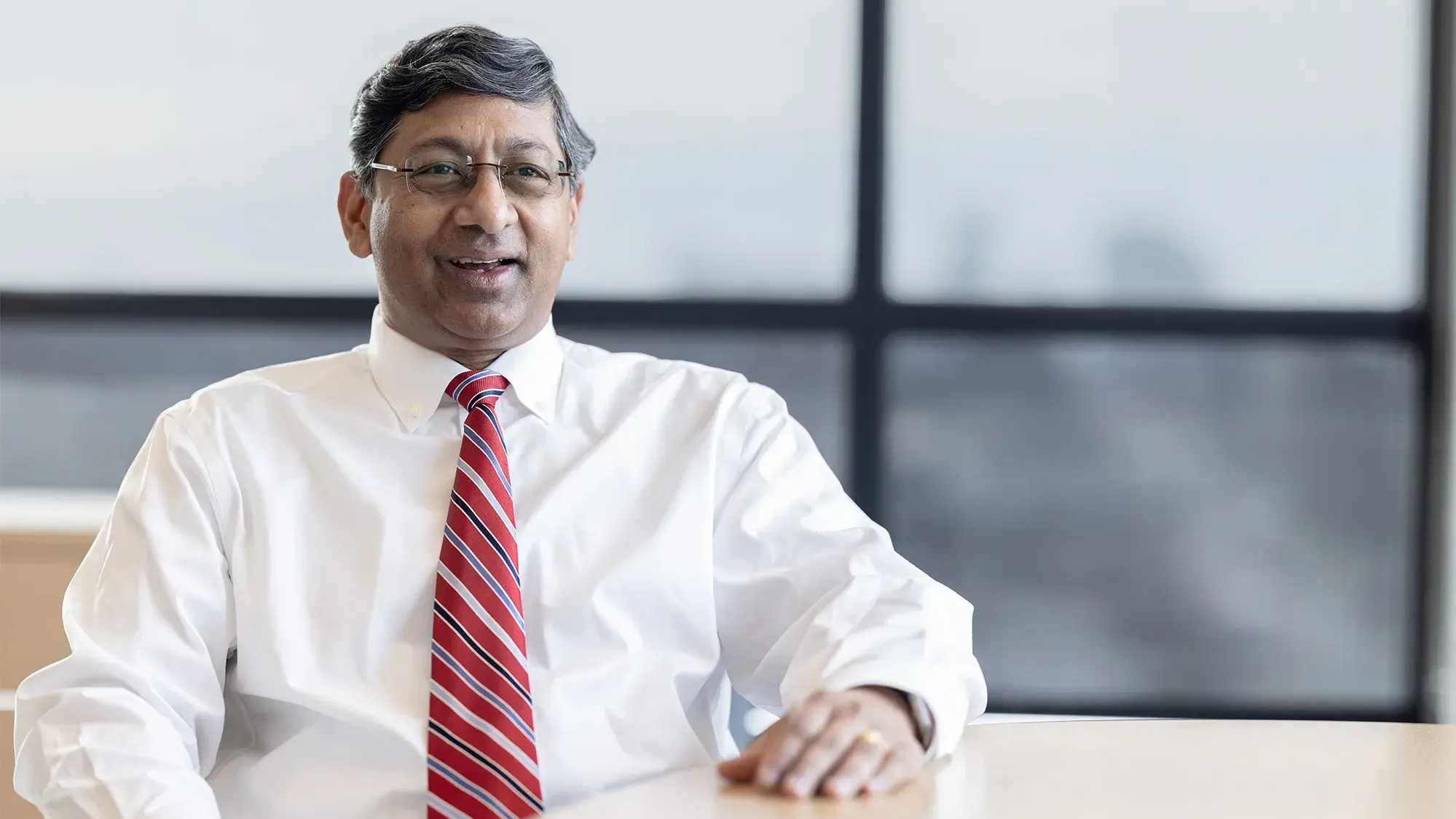 Ravi Bellamkonda sits casually at a table in front of windows overlooking campus. He smiles as he talks.