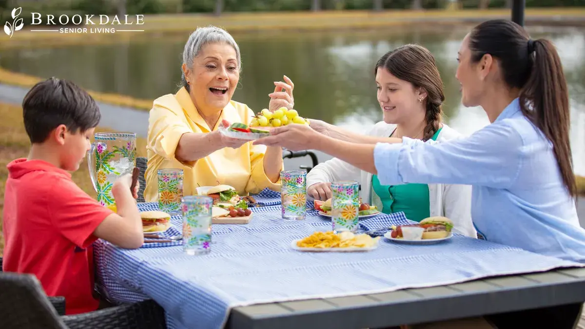 A family sits at an outdoor picnic table by a pond, sharing food and drinks on a blue checkered tablecloth.