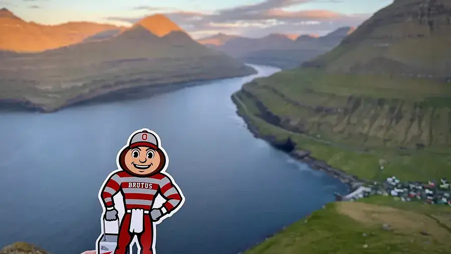 A Brutus Buckeye cutout stands in front of a wide river winding between tall, green mountains at sunset.